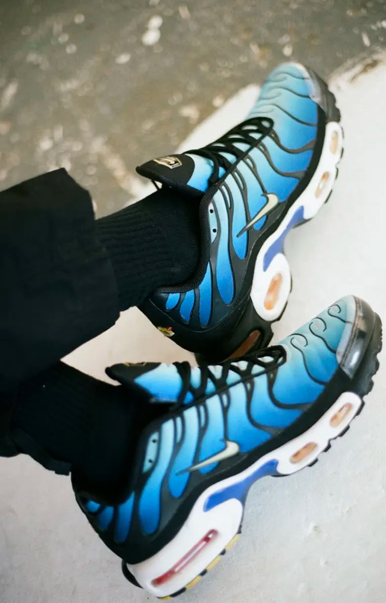 Close‑up view of feet in Nike Air Max Plus TN OG Hyperblue sneakers, with black trouser hems in frame.