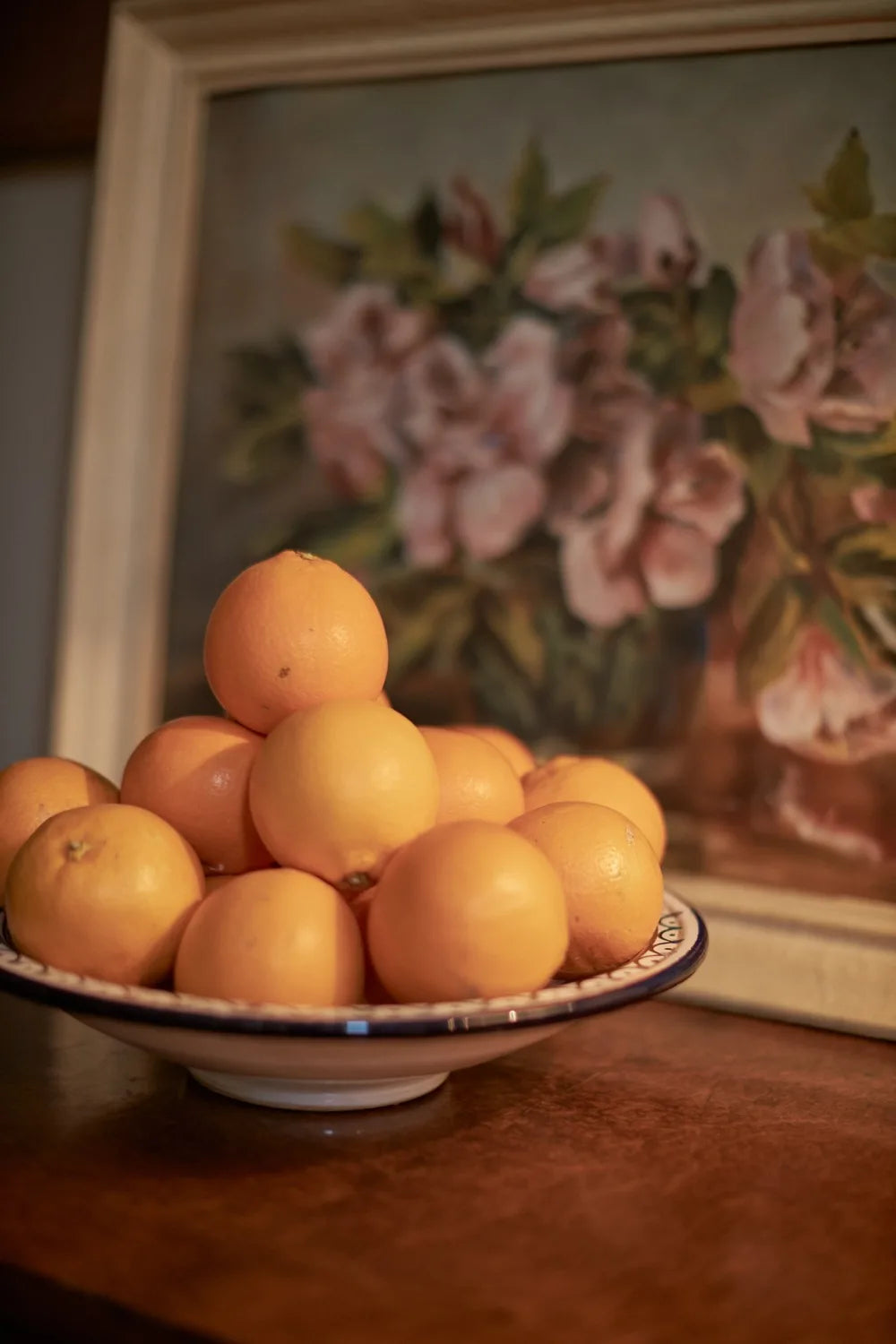 A bowl of oranges in front of a painting of some flowers.