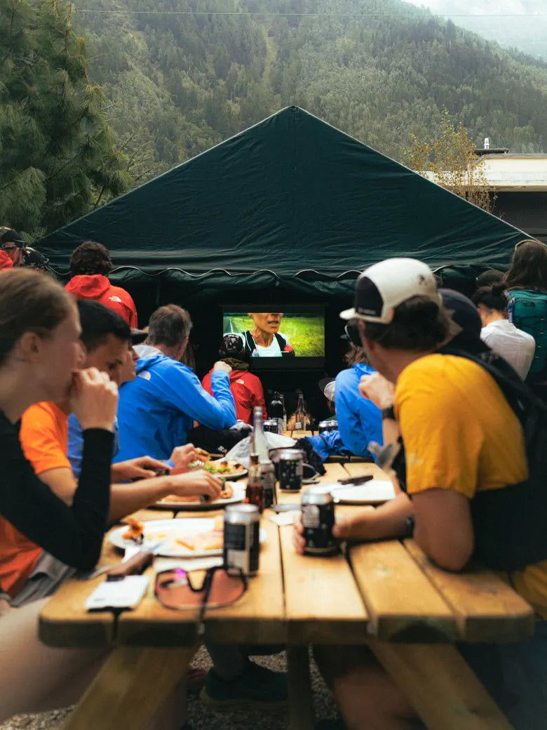 People sat on an picnic bench, eating, watching a screening of a film. A mountain towers in the background.